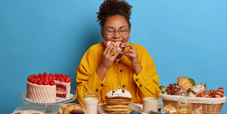 A woman surrounded by sweets takes a bite of a piece of cake.