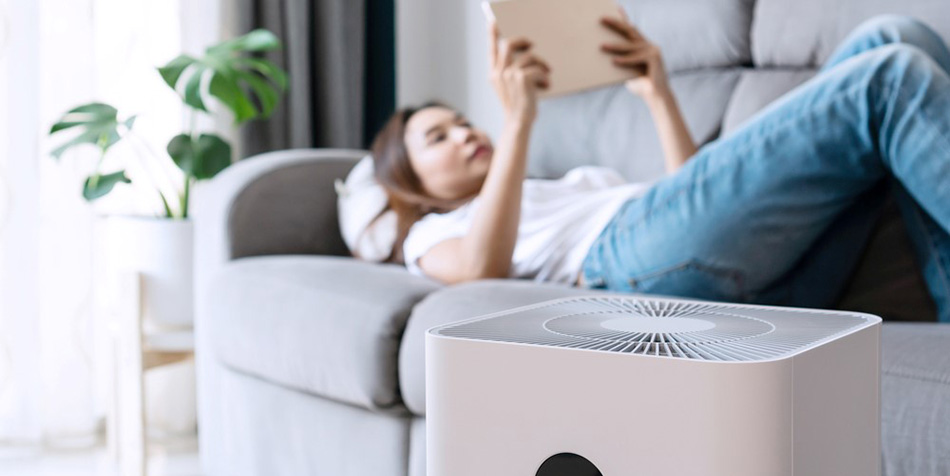 An air purifier runs in the living room while a woman reads her tablet on the couch.